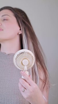 Young woman holding a mini portable fan enjoying cool air and smiling on a hot sunny summer day outdoors.
