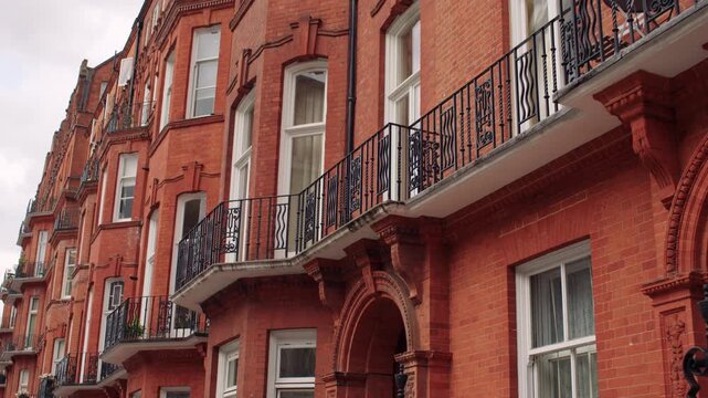 Row of traditional red brick London townhouses with ornate iron railings, arched entrances, and black wrought-iron balconies under a cloudy sky.