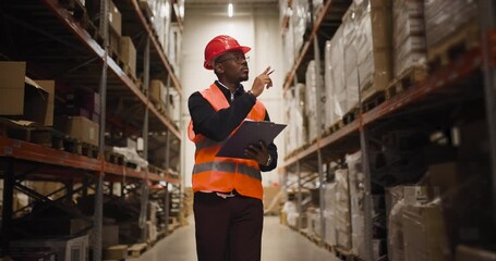 Warehouse worker in safety gear inspecting shelves with a clipboard, ensuring inventory accuracy in a large industrial storage facility - Powered by Adobe