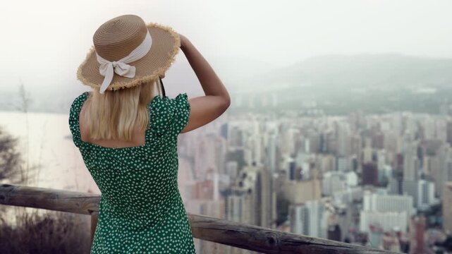 Woman tourist in sundress and floppy hat capturing stunning views of city and ocean with camera from scenic hilltop, relishing holiday and exploration