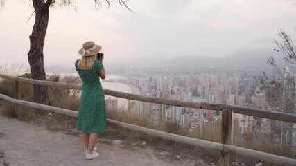 Young woman in straw hat and green dress stands on hill photographing hazy sunset cityscape with beach and skyscrapers, capturing scenic coastal panorama during vacation in Benidorm Spain