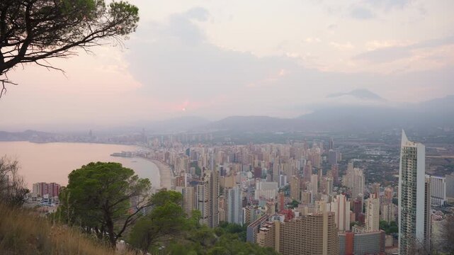 Expansive aerial view of benidorm's cityscape and poniente beach at sunset, showcasing modern skyline and coastal bay with hazy, picturesque sky from  sierra helada viewpoint