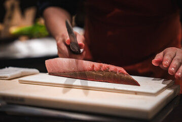 A chef's hands slice a thick tuna fillet with a yanagiba on a white board at a Japanese hotel sushi...