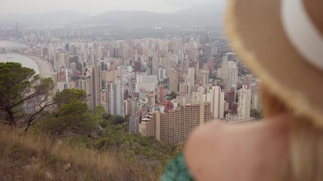 Blonde woman in straw hat admiring stunning cityscape of benidorm from hilltop, taking in summer vacation vibes and picturesque view.Europe travel. Lifestyle, vacation, tourism,nature, active life