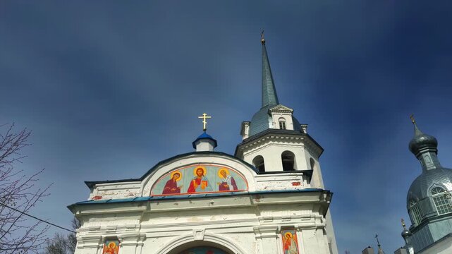 A dramatic low-angle shot of the upper portion of a traditional Russian Orthodox Church, featuring a weathered white facade with a vibrant, semi-circular icon painting of Christ and saints above the
