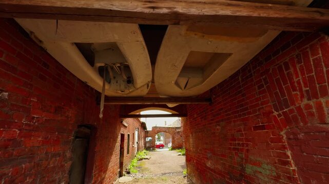 An unusual, dramatic low-angle shot looking through a very narrow, dark passage or archway in an old red brick building, where two small, light-colored boats (likely dinghies or kayaks) are creatively