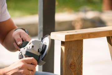 A man's hands sand a homemade wooden bench with a angle grinder outdoor. Close up. DIY