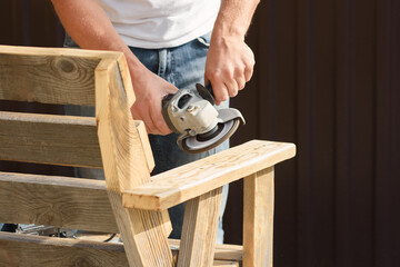 A man's hands sand a homemade wooden bench with a angle grinder outdoor. Close up. DIY