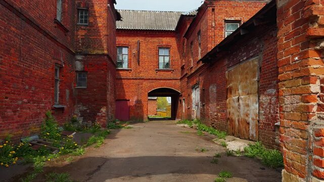 A medium, straight-on shot looking down a narrow, empty dirt and paved courtyard surrounded by tall, aged buildings of deep red brick. The sunlight highlights the weathered texture and peeling stucco