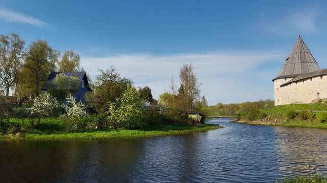 A panoramic, wide-angle shot of a calm river winding past an ancient, towering medieval fortress with light stone walls and a dark conical roof on the right. The left bank contrasts with a picturesque