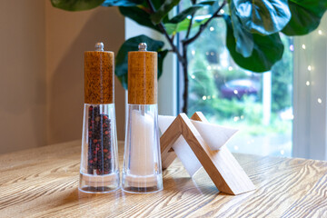 Salt and pepper grinders with wooden tops and napkin holder on wooden table near window with lights. © Anastasiia