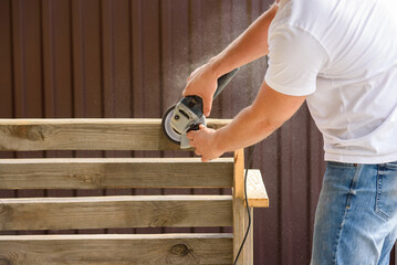 A man sand a homemade wooden bench with a angle grinder outdoor. Close up. DIY