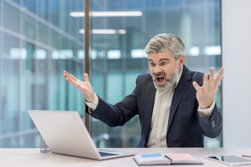 Agitated middle-aged businessman experiencing extreme stress and anger at his desk, reacting to a...