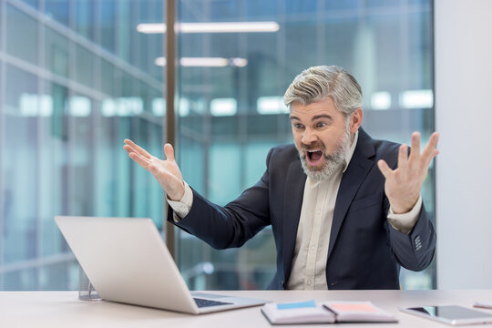 Agitated middle-aged businessman experiencing extreme stress and anger at his desk, reacting to a problem with his laptop in a modern office environment - Powered by Adobe
