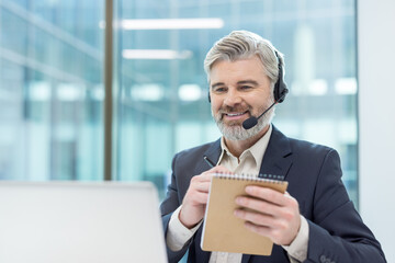 Senior businessman working in call center, smiling while communicating with clients, wearing headset and writing notes, offering customer service and online support
