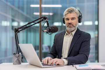 Mature man with headphones and a microphone broadcasting a podcast or online radio show, sitting at a desk and typing on a laptop, creating engaging content