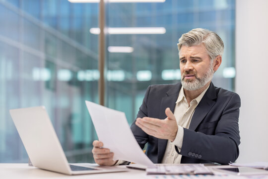Confused senior businessman with white hair studying financial documents at a modern office desk, overwhelmed by paperwork, laptop and budget reports while stressing over finances - Powered by Adobe