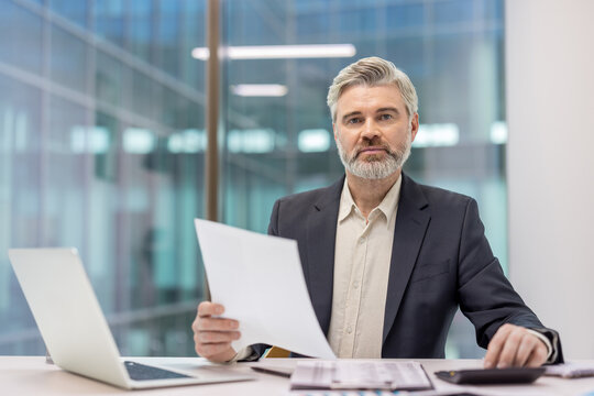 Mature businessman with gray hair and beard working at a modern office desk, reviewing a financial report while using a laptop and calculator, representing finance and executive work