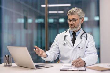 Experienced doctor in a white coat and stethoscope providing an online medical consultation, communicating and gesturing during a telemedicine video call from a modern office using a laptop