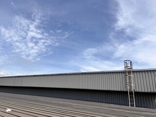 Fototapeta premium A low-angle view of a vast, corrugated metal industrial roof on a factory or warehouse building against a bright blue sky with wispy clouds, featuring a vertical safety cage ladder.
