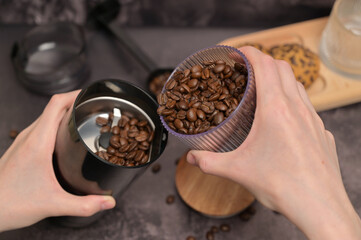 Close up. A woman's hands pours coffee beans into a coffee grinder from a transparent glass. Oatmeal cookies with chocolate chips on a wooden board. Dark background