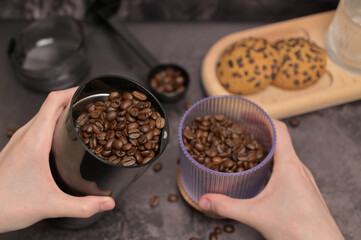 Close up. A woman's hands pours coffee beans into a coffee grinder from a transparent glass. Oatmeal cookies with chocolate chips on a wooden board. Dark background