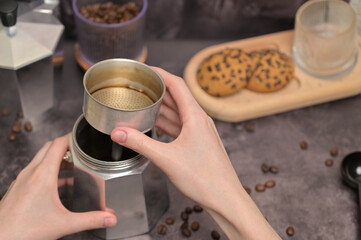 Close up. A woman's hands holding a geyser coffee maker with water. Coffee beans in a glass. Oatmeal cookies with chocolate chips on a wooden board. Dark background