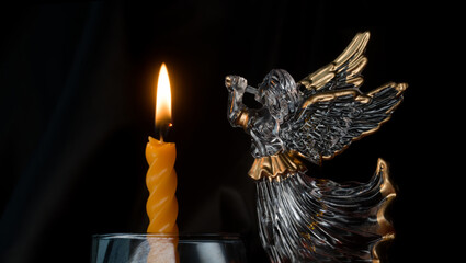 Glass angel with trumpet by a burning spiral candle in a glass on a black backdrop; quiet vigil for the departed, remembrance and prayer in the dark. Photo