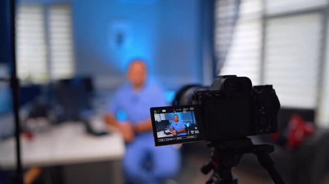Professional male doctor in blue uniform sits ay desk talking to camera. Close up. Medical vlog concept. Selective focus.