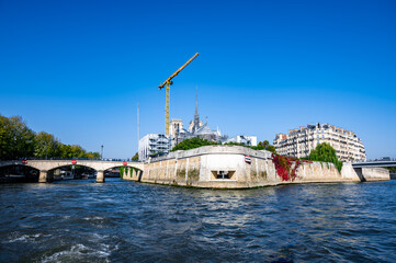 Scenic view of a riverbank with a construction crane, lush greenery, and historic buildings under a clear blue sky, showcasing urban development