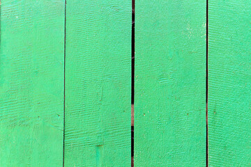 Green painted wooden fence with visible vertical planks and natural wood texture. Surface shows rough grain patterns, small cracks, and gaps between boards, creating rustic outdoor background