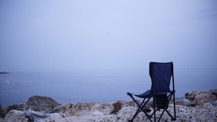A solitary folding chair sits on a rocky shoreline, overlooking a calm, misty ocean.