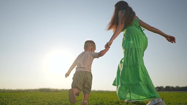 Boy walking in field mother. Woman holds toddler by hand. Outdoor field bright with sun. Toddler steps on grass near mother. Boy walks with woman in grass. Outdoor moment of mother and toddler walking