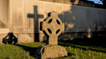 A sunlit, weathered Celtic cross in a graveyard with its strong shadow and the backdrop of a war...