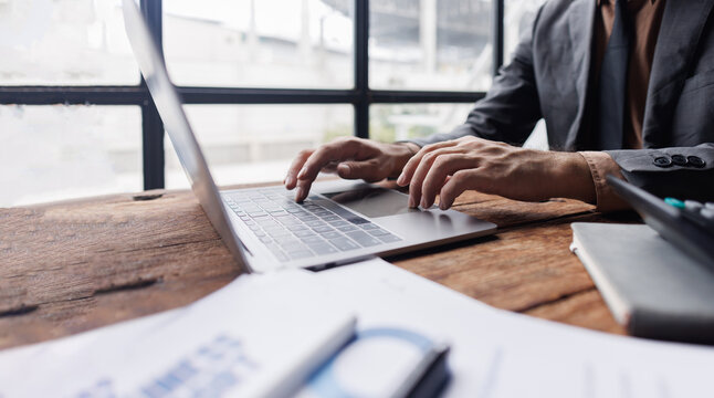 Business man working on laptop computer at modern office. Businessman in black suit working and typing on laptop keyboard, surfing the internet, digital tablet on office desk,