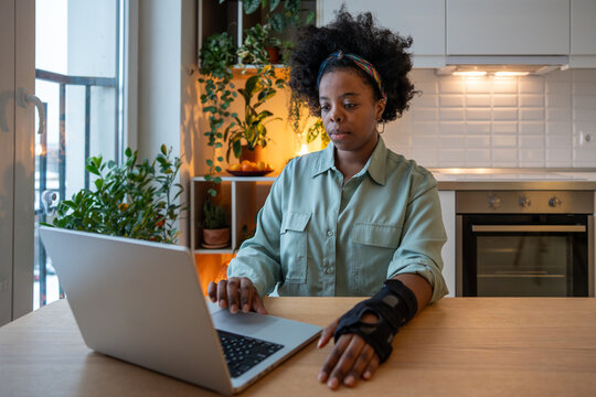 Calm woman with orthopedic wrist brace concentrating on laptop screen. Focused black female freelancer using computer at home kitchen, adjusting to arm injury while working remotely healing process. 