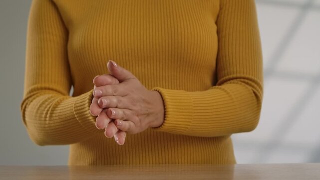 A woman rubs her hands. An unrecognizable woman in a yellow sweater warms her hands in cold weather and rubs them together while standing in a bright room, close-up of her torso and hands. Slow motion