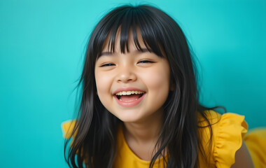A cheerful young child with long dark hair and bangs is laughing joyfully in front of a bright turquoise background.