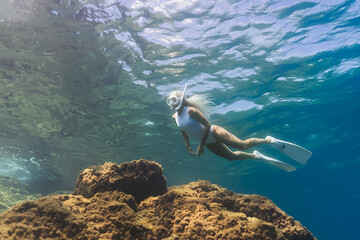 Young woman in a white swimsuit and fins snorkeling in the crystal clear turquoise ocean, exploring the underwater world.