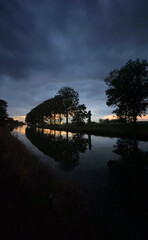 Obraz premium A tranquil river scene at dusk, showcasing the beauty of silhouetted trees against dramatic clouds overhead
