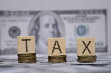 Wooden blocks spelling TAX on stacks of coins with US dollar bills in the background symbolize taxation, financial responsibility, accounting, and government revenue collection.