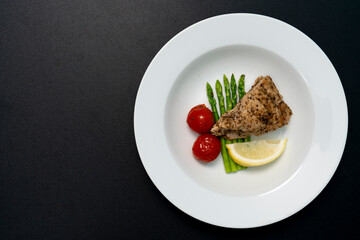 Red snapper steak, asparagus, tomatoes, and lemon on a white plate on a black background. Table top view.
