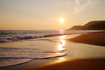 A serene sunset casts warm, golden light over a quiet beach. Gentle waves wash the sand while a distant hillside rises beyond a simple pier, creating a peaceful coastal landscape.