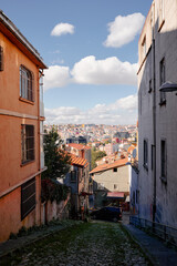 A narrow cobblestone street runs between orange and gray buildings, guiding viewers toward a sunlit cityscape of terracotta roofs and hillside apartments under a clear blue sky.