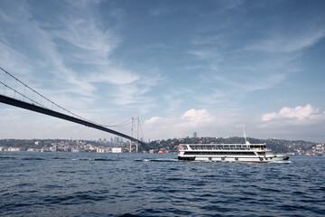 A picturesque view of a ferry floating on a large body of water beneath a bridge, amidst a tranquil coastal city setting. The clear sky complements the pleasant and serene atmosphere.
