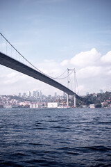 Image features a large suspension  Istanbulbridge crossing a river, with an urban cityscape and clear skies extending into the horizon.