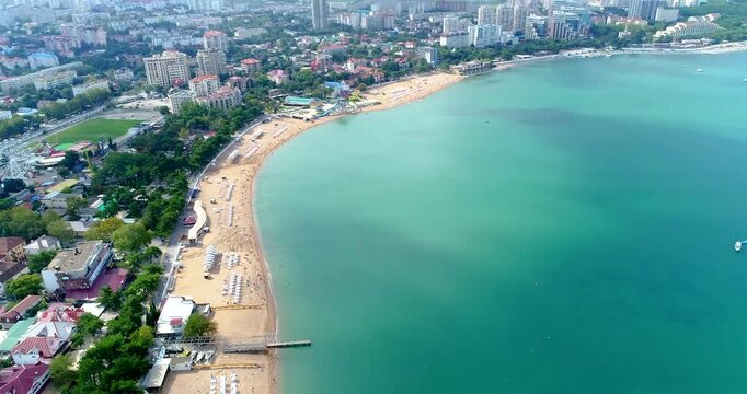 Gelendzhik resort. Flight over central beach towards the "Tolstoy" Cape. On beach there are sun loungers, umbrellas, people sunbathing. Promenade along beach. Beautiful emerald sea, sun