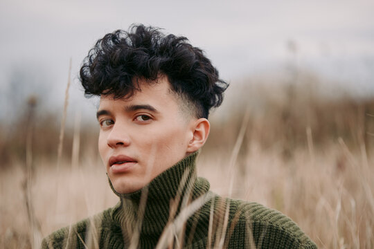 A close portrait of a young person in tall dry grass wearing a textured sweater, conveying quiet confidence, authenticity and credible presence through natural light and unposed expression.