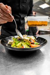Chef drizzling dressing over fresh salad in black bowl, in professional kitchen. Ingredients include lettuce, tomato, olives, and feta. Culinary preparation in restaurant setting.
