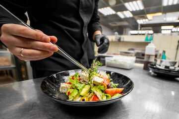 Chef in black gloves garnishing fresh salad with metal tongs in professional kitchen. Bowl contains feta, avocado, cherry tomatoes, lettuce, herbs. Culinary preparation concept.